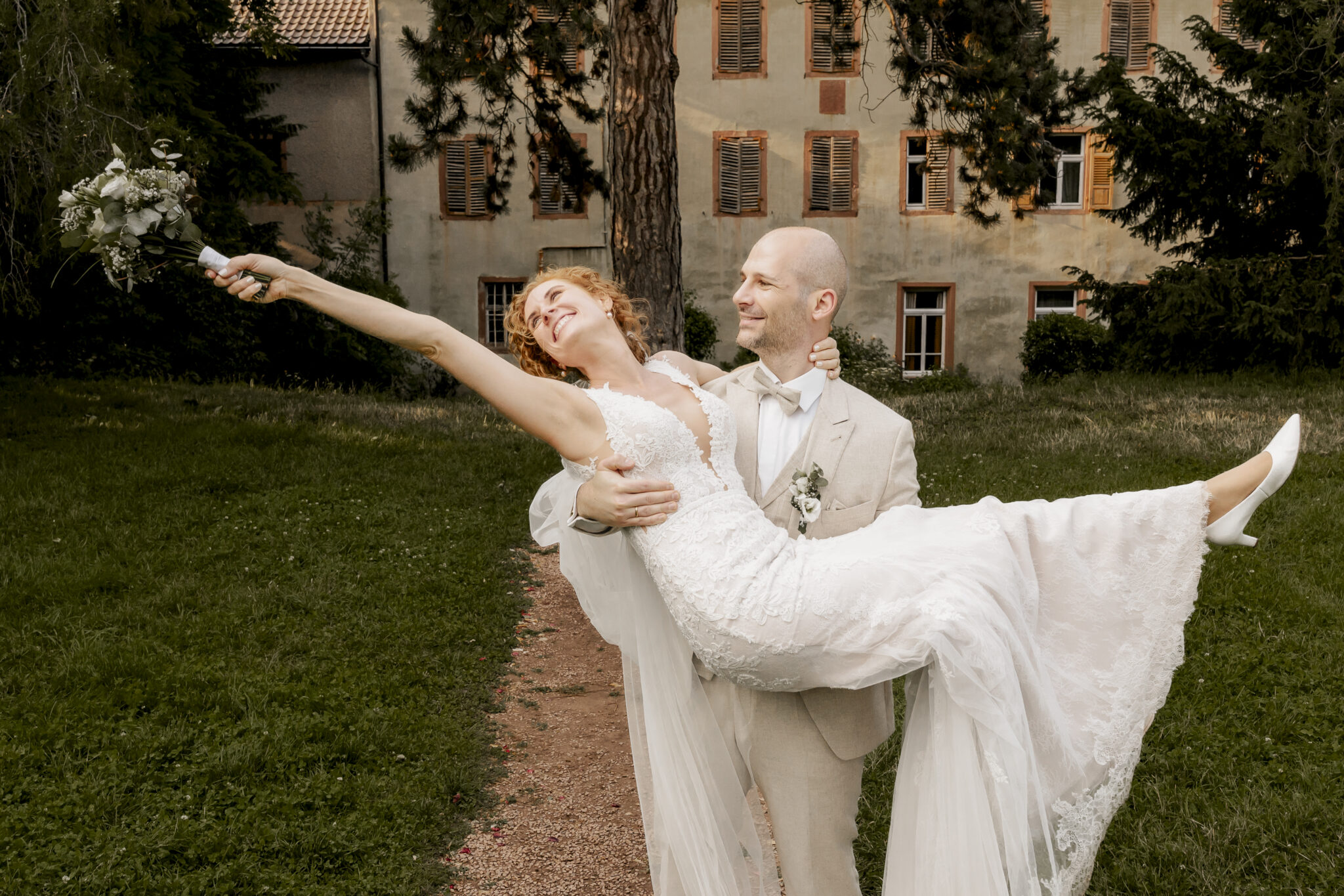 Bräutigam trägt die Braut auf einem Gartenweg, während sie ihren Blumenstrauß seitlich ausstreckt.