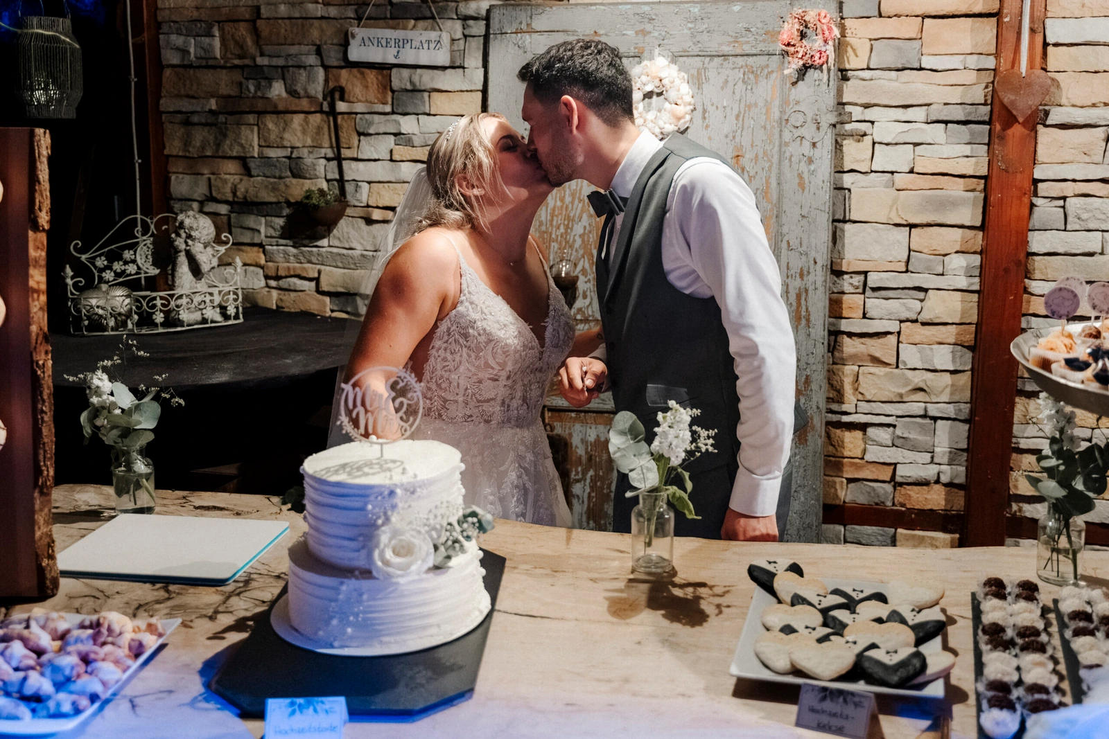 Laura und Philipp küssen sich neben ihrer zweistöckigen Hochzeitstorte auf einem Tisch.